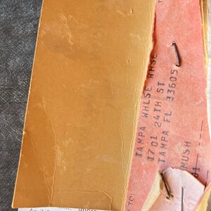 Puede incluir: Primer plano de ef&iacute;meras de papel vintage, que incluyen una etiqueta naranja rasgada y un boleto rosa. El texto incluye "TAMPA WHLSE WHSE" y "American of Martinsville". El papel est&aacute; envejecido y grapado.
