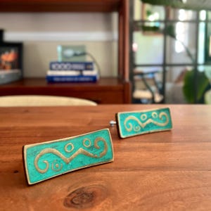 May include: Two rectangular cabinet knobs featuring a vibrant turquoise finish with an inlaid gold-toned metal design. The metal pattern consists of a central horizontal line with curved ends and small circular accents, creating a distinct visual contrast.