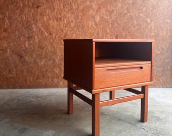 May include: A mid-century modern nightstand in a warm reddish-brown hue. The wooden bedside table features a single drawer with a recessed handle and an open shelf. The table stands on four slender legs, showcasing a minimalist design.