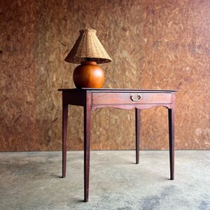 May include: A wooden side table with a single drawer featuring a brass handle, topped with a round wooden lamp base and a conical woven wicker lampshade. The table has four straight legs and a curved apron detail beneath the drawer front.