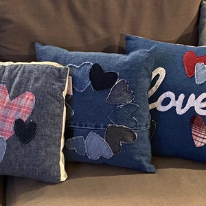 May include: Four denim pillows with different heart designs. One pillow has a pink, red, and brown heart, another has a circle of blue hearts, another has a red and blue heart, and the last one has the word "Love" in white with a plaid heart.