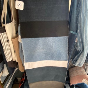 Op de afbeelding: Een spijkerbroekrok met een patchwork-ontwerp, met verschillende tinten blauwe denim, een zwarte band en een witte streep. De rok hangt aan een metalen hanger en toont zijn unieke, handgemaakte stijl. De rok is ongeveer 76 cm lang.