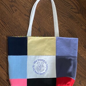 May include: A patchwork tote bag with a white strap and a peace sign embroidered on the front. The bag is made of different colored fabrics, including blue, yellow, pink, red, and black. The embroidery says "The world is better with you in it".