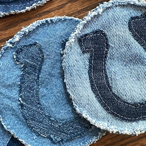 May include: Four blue denim coasters with frayed edges and a dark blue horseshoe design.