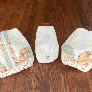 May include: Three quilted zippered pouches with floral patterns in shades of green, red, and yellow. The pouches are lined up in a row on a wooden surface.