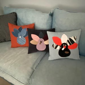 May include: Four decorative throw pillows with a Thanksgiving theme. Each pillow features a different design of a turkey with colorful fabric accents. The pillows are arranged on a light blue couch.