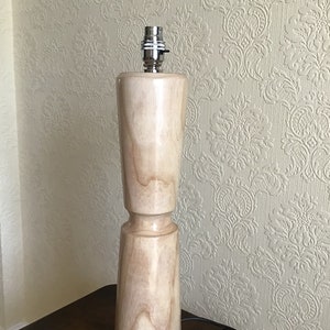 May include: A wooden table lamp with a natural wood finish. The lamp has a simple, cylindrical design with a slightly flared base. The lamp is turned on and the light is off.