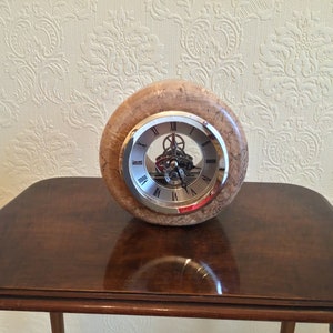 May include: A round clock with a light brown, speckled wooden casing. The clock face has a silver rim and Roman numerals. The clock is displayed on a dark brown wooden table.