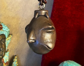 May include: A bronze-toned pendant shaped like a rounded head with sculpted facial features including a nose, closed eyes, and a mouth. The pendant is suspended from a dark brown leather cord by a metal loop and a knot at the top of the object.