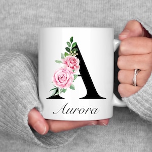 May include: White ceramic mug featuring a black letter A and a pink floral design. The mug has the name "Aurora" printed beneath the letter.