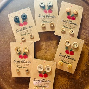 May include: A collection of button earrings displayed on brown cards. Each card features two button earrings and smaller accent buttons. The cards are labeled "Sweet Blunder" with the text "Where vintage meets new life. Thank you!"
