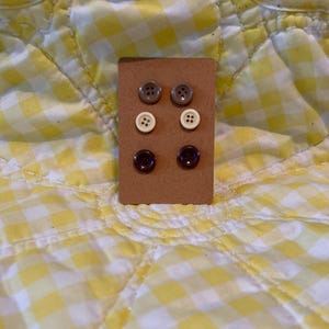 May include: A set of six button earrings on a brown card. The earrings are in three different colors: brown, white, and beige.
