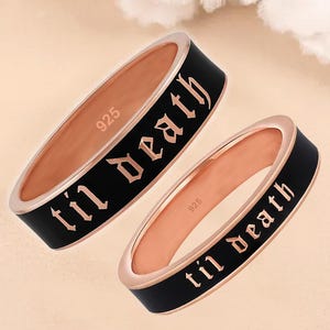 May include: Two rose gold rings with black enamel bands. The rings have the words "til death" in gothic font. The inside of the rings are stamped with "925". The rings are displayed on a light beige background.