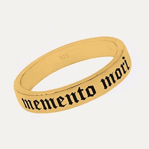 May include: A gold-colored ring with the inscription "memento mori" in black gothic font. The ring is a simple band design, and the interior is stamped with "925". The ring is likely made of sterling silver with a gold finish.