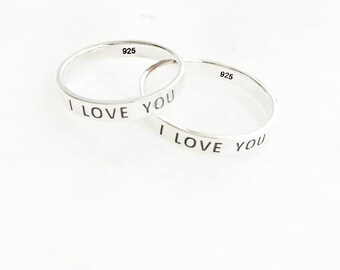May include: Two silver rings with the inscription "I LOVE YOU" in black lettering. Each ring is stamped with "925". The rings are displayed on a white surface, showcasing a simple and elegant design.