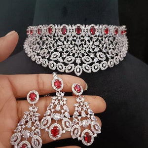 May include: A silver choker necklace with red stones and matching dangle earrings. The necklace is made of silver and features a floral design. The earrings are also silver and feature a floral design with red stones.