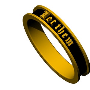 Può includere: Un anello color oro con una fascia nera con la scritta "Let them" in un carattere gotico. L'anello ha un design arrotondato e liscio.