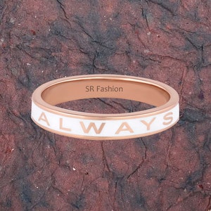 May include: A rose gold ring with a white enamel band that says "ALWAYS" in white lettering.
