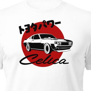 Può includere: Maglietta bianca con un'illustrazione in bianco e nero di un'auto Toyota Celica davanti a un cerchio rosso con il testo giapponese "トヨタパワー" e la parola "Celica" sotto.