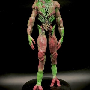 May include: A 3D printed figurine of a humanoid alien with a small alien on its shoulder. The alien is green and pink with a detailed, textured surface.