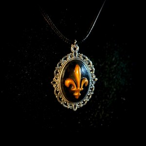 Peut inclure: Un collier pendentif avec un pendentif ovale noir orné d'un motif de fleur de lys doré. Le pendentif est serti dans un cadre filigrané de couleur argentée et est suspendu à un cordon noir. Le fond est noir.