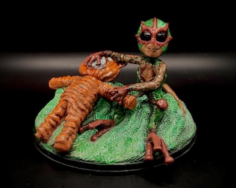May include: A handcrafted figurine depicting a creature with a green and red mask, holding a figure with orange stripes. The figures are on a green base with a black border. The overall theme is fantasy.