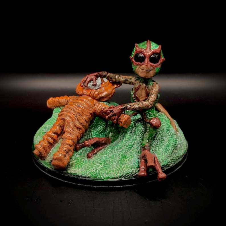 May include: A handcrafted figurine depicting a creature with a green and red mask, holding a figure with orange stripes. The figures are on a green base with a black border. The overall theme is fantasy.