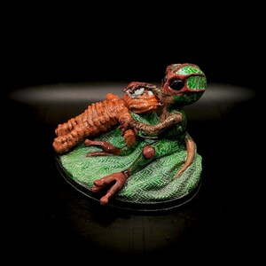 May include: A whimsical sculpture of a green and red alien figure holding an orange creature. The alien is seated on a green patterned base. The sculpture is set against a black background.