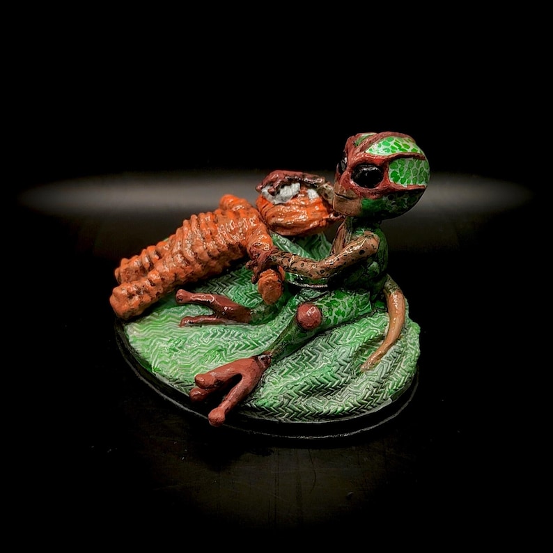 May include: A whimsical sculpture of a green and red alien figure holding an orange creature. The alien is seated on a green patterned base. The sculpture is set against a black background.