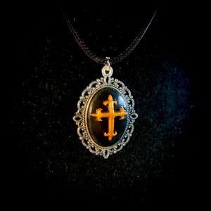 Peut inclure: Collier pendentif ovale avec une croix dorée sur fond noir, encadrée d'une monture en filigrane argenté. Le pendentif est suspendu à un cordon noir. Un accessoire d'inspiration gothique.