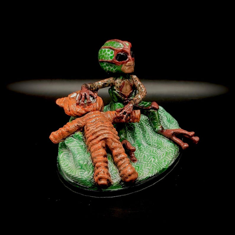 May include: A detailed figurine of an alien figure with a green and red patterned head, seated on a green patterned base, interacting with a smaller orange figure. The alien has brown and red skin tones.
