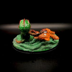 May include: A miniature sculpture featuring a green, spotted creature with a red-trimmed head, seemingly interacting with an orange, textured form. The figures rest on a textured green base with a black border, set against a dark background.