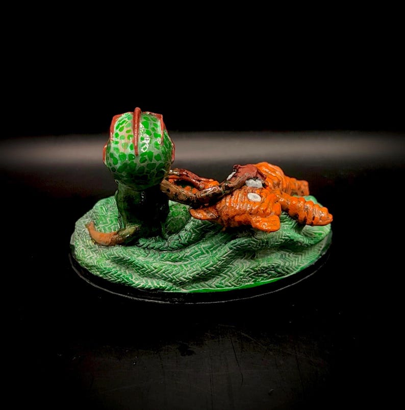 May include: A miniature sculpture featuring a green, spotted creature with a red-trimmed head, seemingly interacting with an orange, textured form. The figures rest on a textured green base with a black border, set against a dark background.