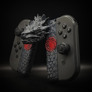Peut inclure: Une manette de jeu Nintendo Switch noire avec une tête de dragon sur le dessus et un sceau rouge sur le côté. La manette a un design texturé et écailleux.
