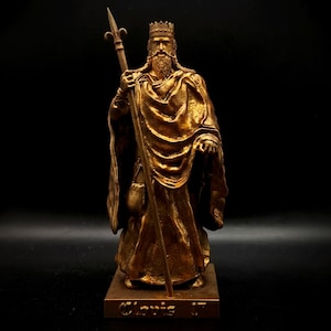 Puede incluir: Estatua de color bronce de un rey con corona y sosteniendo un cetro con un remate de flor de lis. La estatua tiene una larga barba y está cubierta con una túnica fluida. La base de la estatua está inscrita con el nombre "Clovis 1".