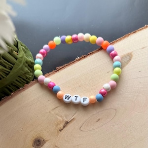 May include: A colorful beaded bracelet with the letters "WTF" spelled out in white beads. The bracelet features a variety of pastel and bright-colored beads, including pink, yellow, orange, and blue. The bracelet is displayed on a wooden surface.