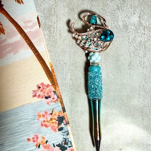 May include: A decorative pen with a teal and gold barrel, topped with a sparkling embellishment. The embellishment features a teal jewel, clear rhinestones, and a gold-colored swan design. The pen is set against a textured gray background, with a floral and palm tree design in the background.