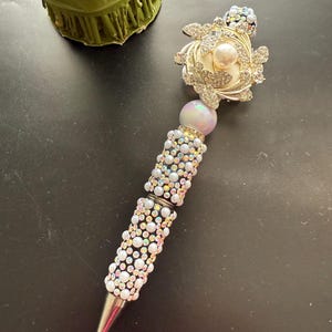 May include: A decorative pen with a silver tip and a barrel covered in iridescent faux pearls and rhinestones. The pen features a large pearl at the top and a floral design with rhinestones and a pearl accent. The pen is a unique writing accessory.