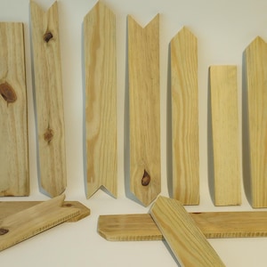 May include: A set of 12 unfinished wooden fence pickets with pointed tops. The pickets are arranged in a row, with some lying on their sides. The pickets are all the same size and shape, and they are made of a light-colored wood.