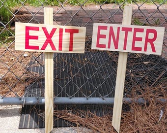 May include: Two rectangular light-colored wooden signs mounted on vertical wooden stakes. The left sign features the word EXIT in bold red capital letters, and the right sign features the word ENTER in bold red capital letters, both with a natural wood grain finish.