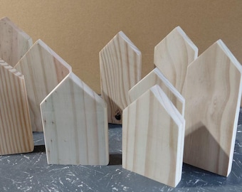 May include: A collection of nine unfinished wooden house-shaped blocks with visible natural wood grain patterns. Each piece features a simple peaked roof design and a flat base, showcasing the light tan color and texture of the raw pine wood material.