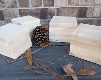 May include: Four stacks of unfinished wooden squares, each stack is about 4 inches tall. The squares are arranged on a dark gray surface with a pine cone and dried leaves.
