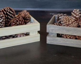 May include: Two rectangular wooden crates filled with brown pinecones. One crate is made of light, natural-colored wood, while the other crate is finished in a weathered gray stain. Both crates feature an open-slat design on the sides.