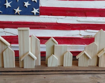 May include: A collection of small, unfinished wooden house-shaped blocks arranged in front of a weathered American flag. The houses vary in size and design, with some featuring pitched roofs.