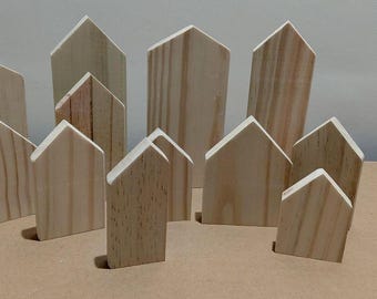 May include: A collection of unfinished wooden house-shaped blocks. The blocks vary in size and are arranged to resemble a small town. The natural wood grain is visible, and the blocks are unpainted.