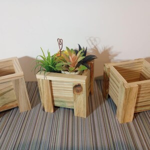 May include: Three wooden planters of varying sizes. The planters are made of light-colored wood and have a square shape. One planter contains a small succulent arrangement with green and orange leaves. The planters are on a striped surface.