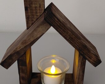 May include: A house-shaped candle holder crafted from dark stained wood, featuring a peaked roof and a chimney. A clear glass votive cup sits at the base, containing a white battery-operated tea light with a glowing yellow flame.