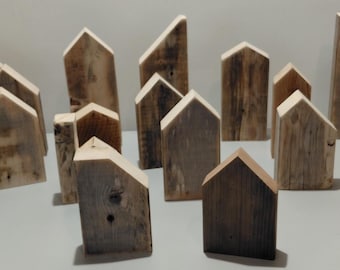 May include: A collection of small, house-shaped wooden blocks crafted from reclaimed wood. Each piece features a unique grain pattern, natural wood tones ranging from light tan to dark gray, and a distinct roofline, including gabled and slanted tops.