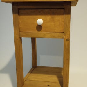 May include: A small, wooden nightstand with a single drawer and a lower shelf. The nightstand is constructed from light-colored wood and features a white, round drawer pull. The dimensions are approximately 24 inches high, 16 inches wide, and 12 inches deep.