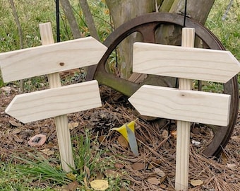 May include: Two light-colored wooden directional signs, each featuring two blank arrow-shaped boards mounted on a vertical post. The natural wood grain is visible on the smooth surfaces of the four horizontal arrows and the two supporting posts.